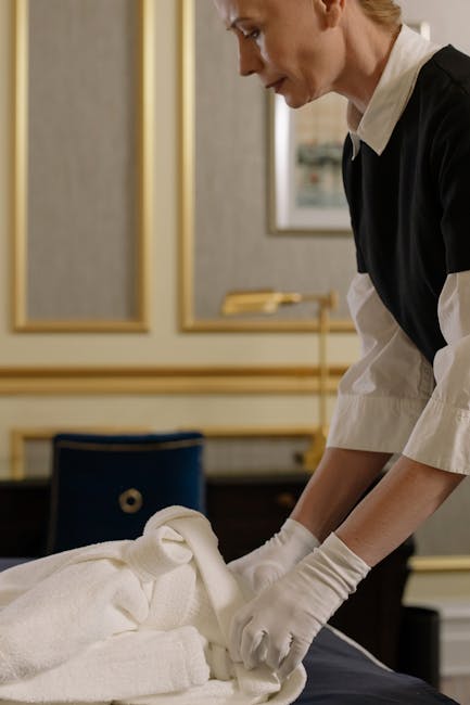 A professional cleaner from Hammersmith Carpet Cleaners, dressed in black and white uniform with gloves, is carefully folding and arranging white towels on a dark-colored table in a well-lit, elegant 