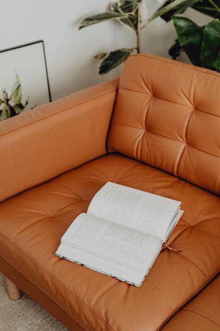 Close-up of a hand holding a vacuum cleaner nozzle, actively cleaning a green upholstered armchair, with a dark gray sofa visible in the background, in a well-lit living room setting, highlighting the